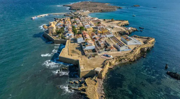 Vista aérea de una pintoresca cala del sur, con casas blancas y tejados naranjas, rodeada por el mar azul y una fortaleza.