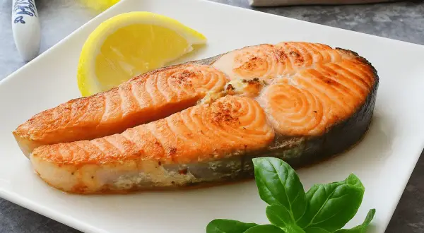 Un jugoso salmón a la plancha, servido con una rodaja de limón y hojas de albahaca fresca en un plato blanco.