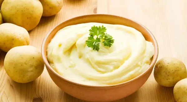 Un cremoso pure de patatas, decorado con perejil, junto a patatas frescas sobre una mesa de madera.