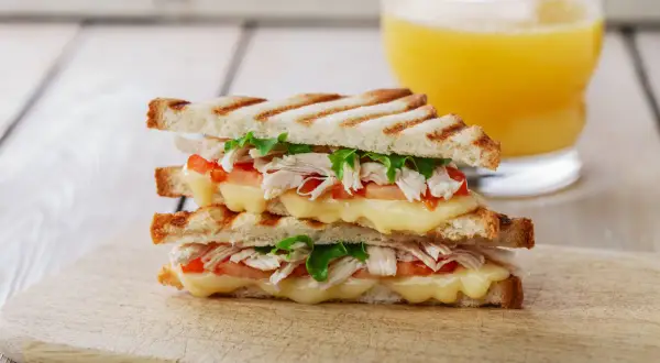 Delicioso sandwich de pollo a la parrilla con queso derretido, tomate y lechuga, acompañado de un vaso de jugo de naranja.
