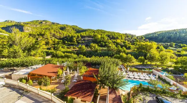 Un día soleado en la rosaleda de Mijares, con un complejo turístico y piscina rodeado de exuberante vegetación y montañas verdes.
