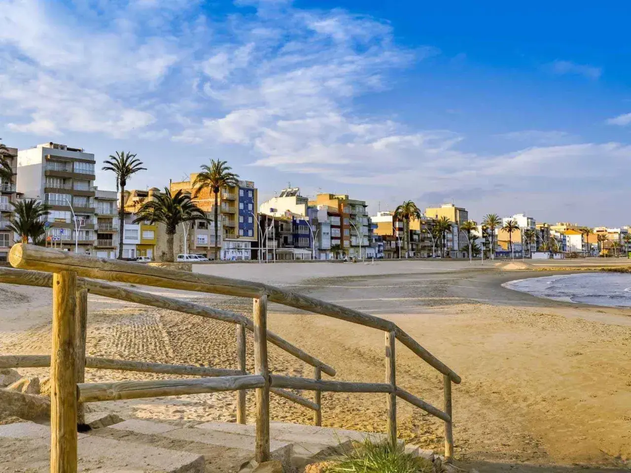 Escaleras de madera bajan a una playa vacía con edificios coloridos y palmeras al fondo. Un ejemplo de pueblos bonitos cerca de Benicàssim.