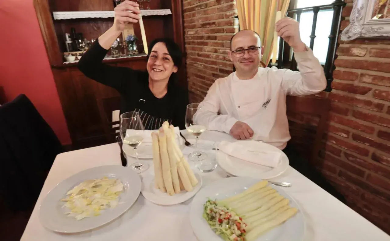 Pareja sonríe mostrando espárragos blancos, uno de ellos en un plato con ensalada y el otro en un manojo atado.