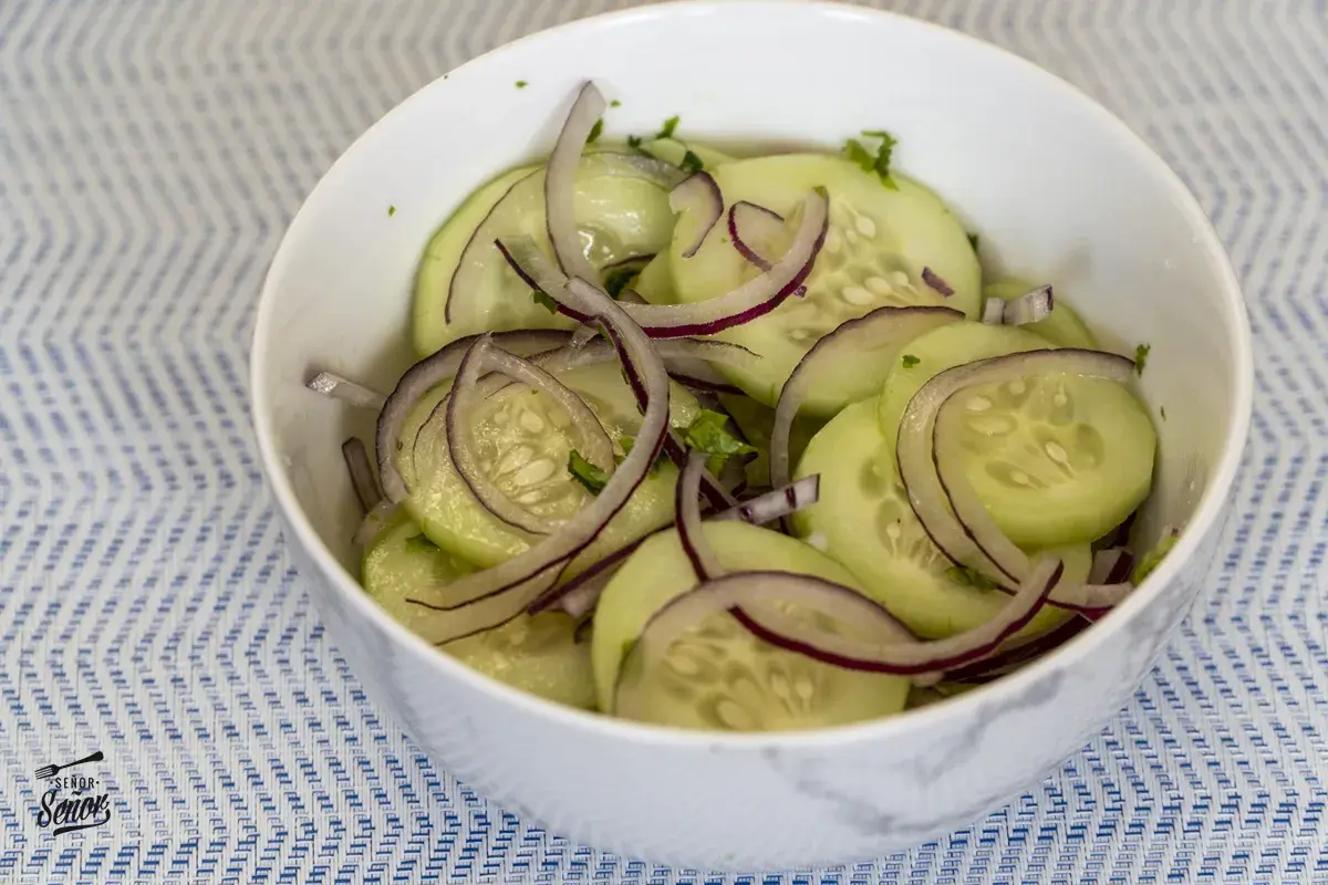 Deliciosa ensalada de pepino con cebolla morada en un tazón blanco sobre un mantel azul y blanco.