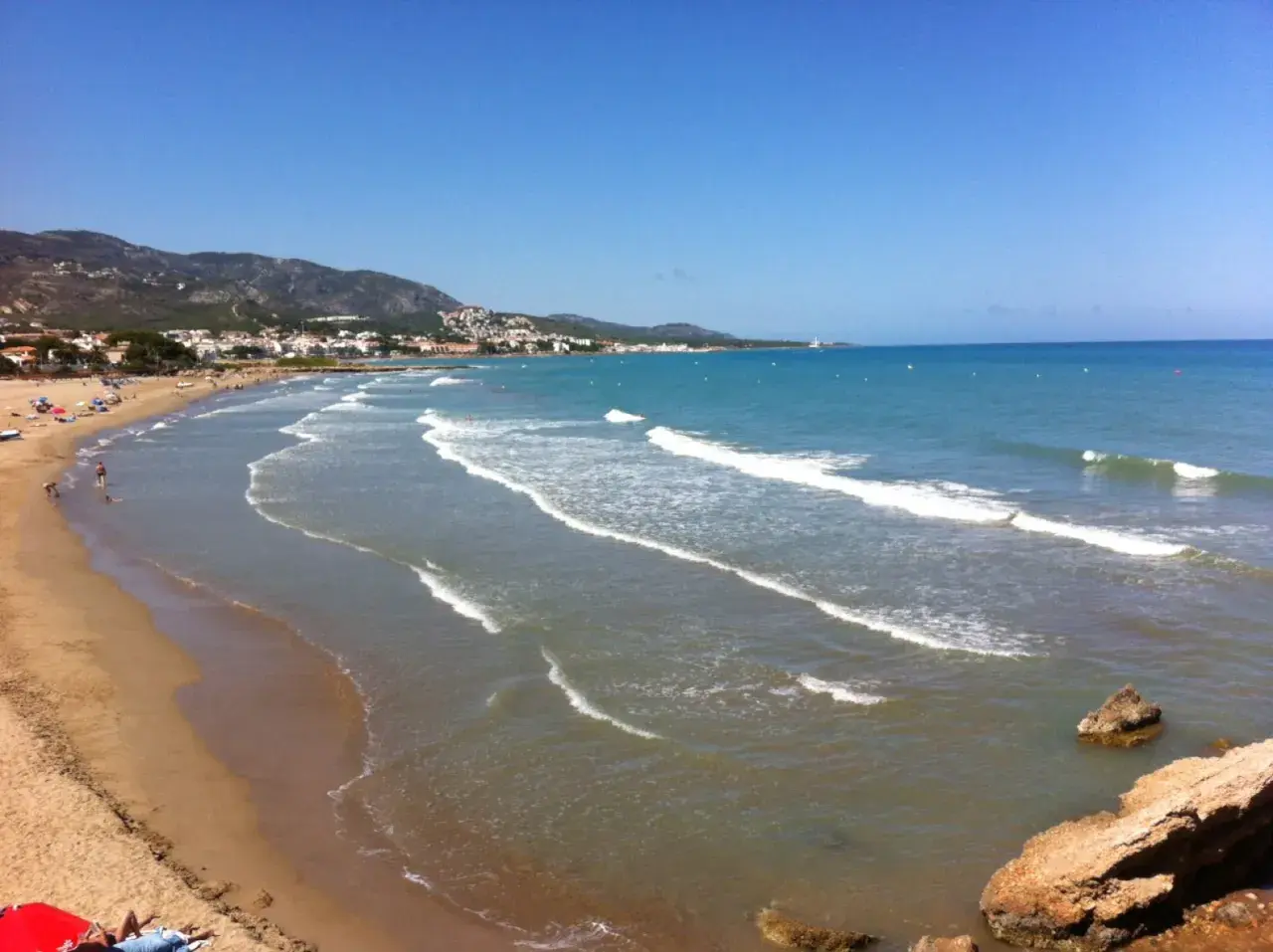 La **playa romana alcossebre** se extiende con arena dorada y aguas turquesas bajo un cielo azul. Olas suaves rompen en la orilla, mientras la costa se dibuja con casas y montañas al fondo.