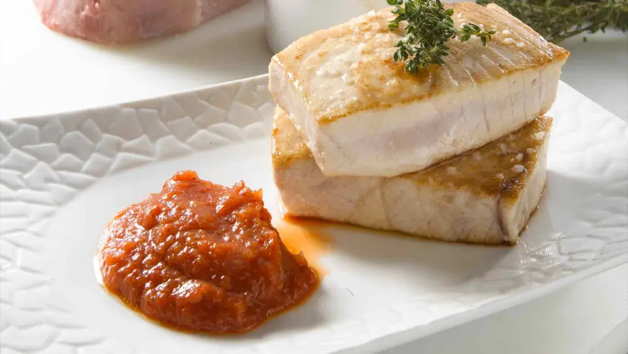 Dos trozos de atún a la plancha apilados, con una guarnición de salsa de tomate y una ramita de tomillo.