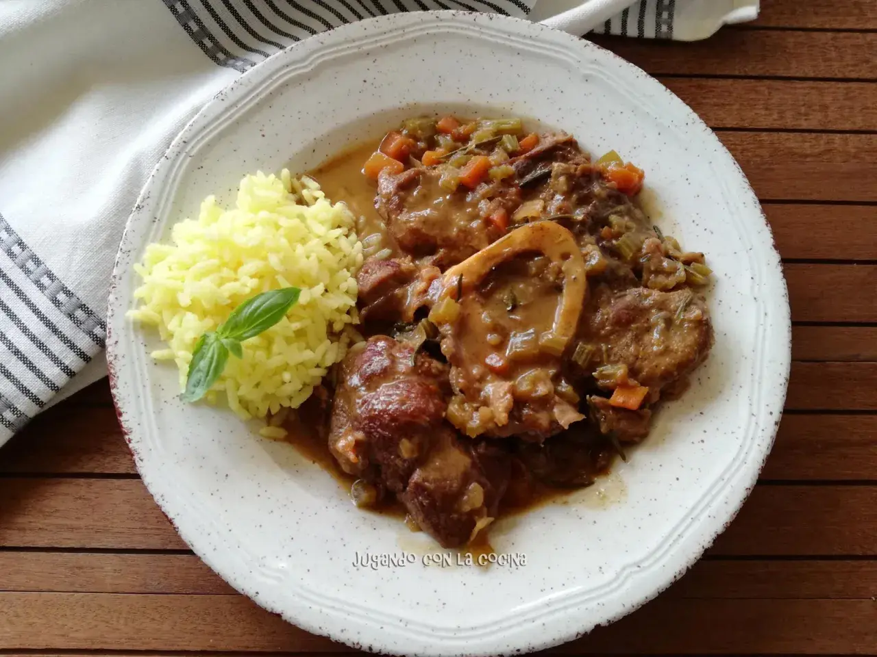 Un plato de osso buco con arroz amarillo y una hoja de albahaca. La carne está cubierta de una salsa con verduras.