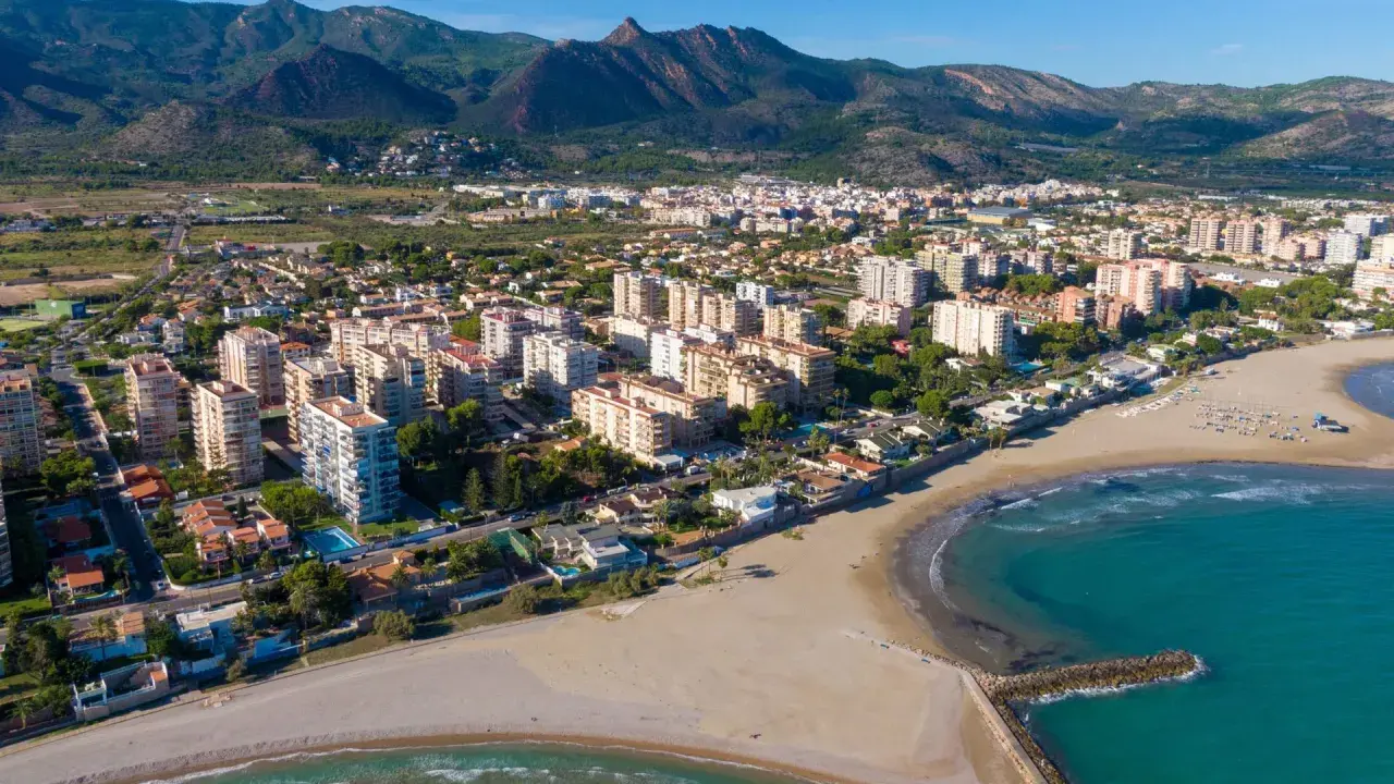 Vista aérea del casco antiguo de Benicàssim pueblo, con edificios residenciales, playas de arena y el mar Mediterráneo. Montañas verdes al fondo.