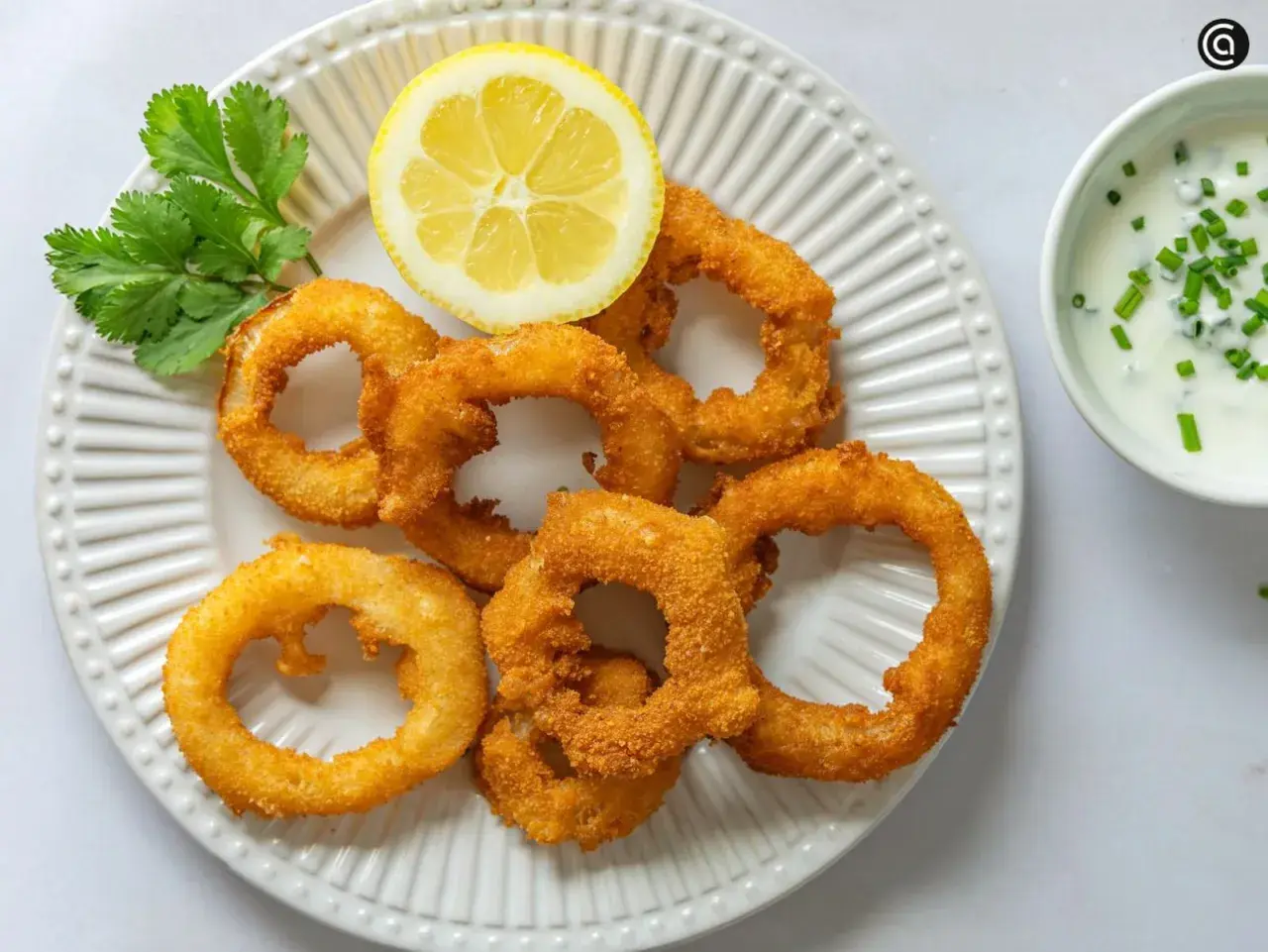 Crujientes aros de cebolla dorados, servidos con una rodaja de limón y un dip cremoso. ¡Un aperitivo irresistible!