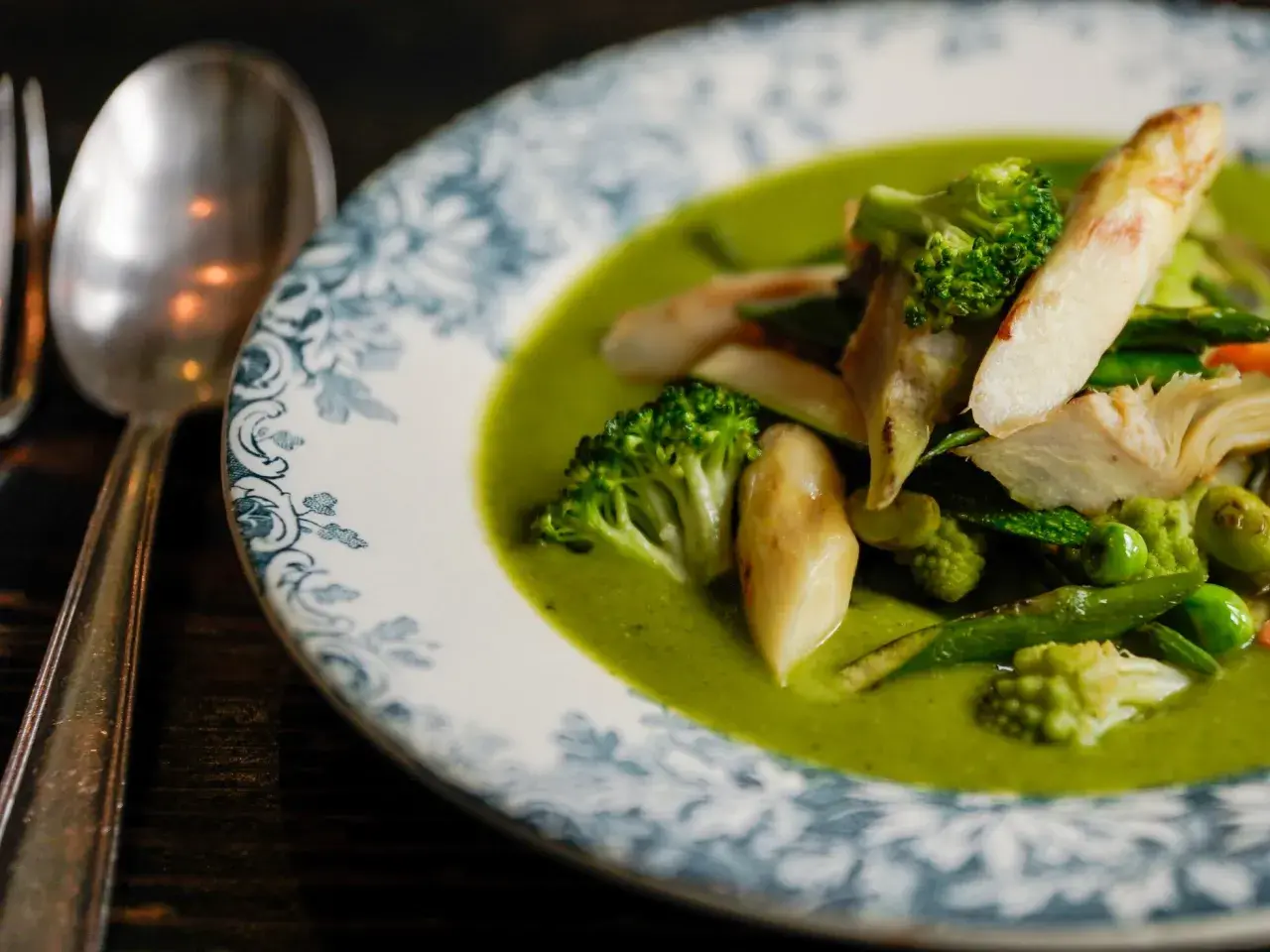 Deliciosa menestra de verduras en un plato con borde azul y blanco. Brócoli, espárragos y guisantes flotan en una cremosa salsa verde.