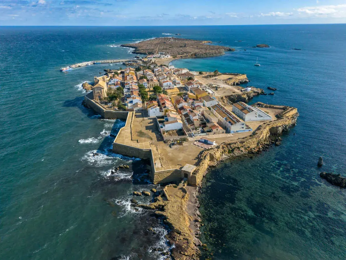 Vista aérea de una pintoresca cala del sur, con casas blancas y tejados naranjas, rodeada por el mar azul y una fortaleza.