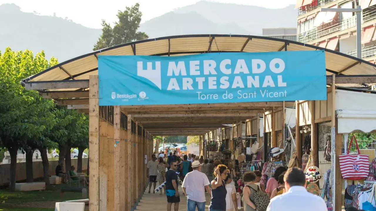 Gente pasea por un mercadillo en Benicàssim, bajo un cartel azul que anuncia "Mercado Artesanal Torre de Sant Vicent".