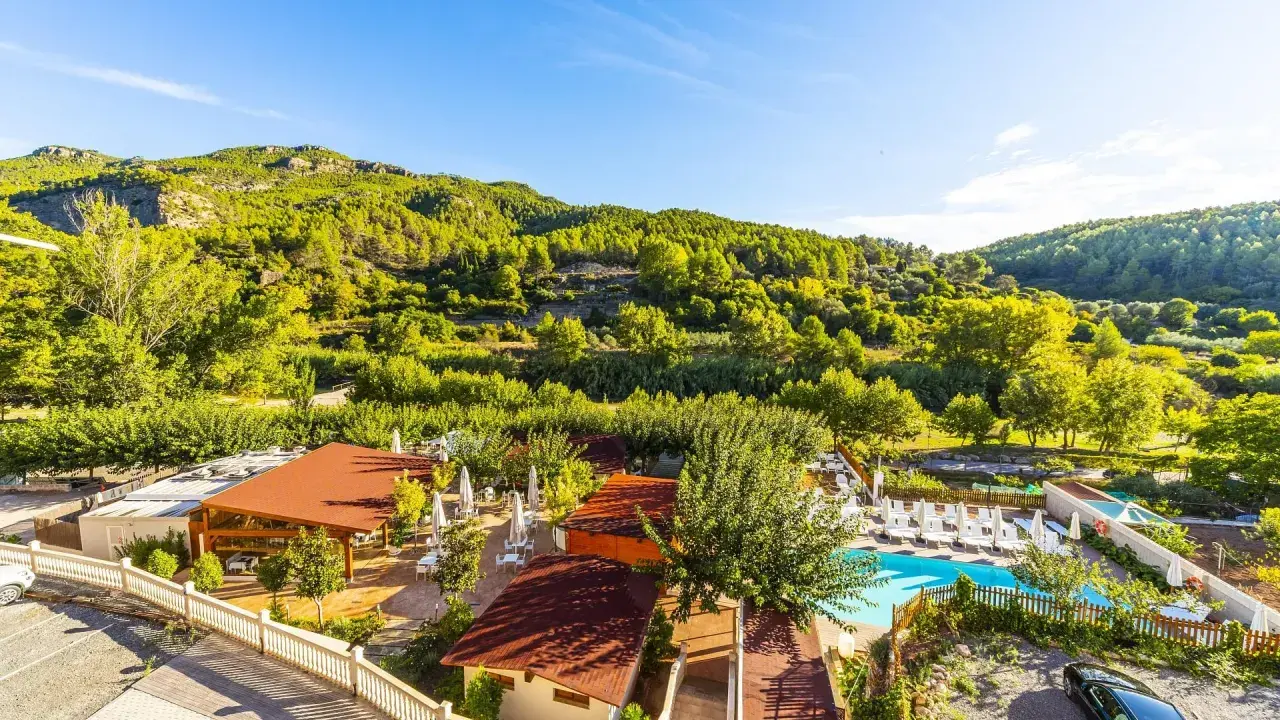 Un día soleado en la rosaleda de Mijares, con un complejo turístico y piscina rodeado de exuberante vegetación y montañas verdes.