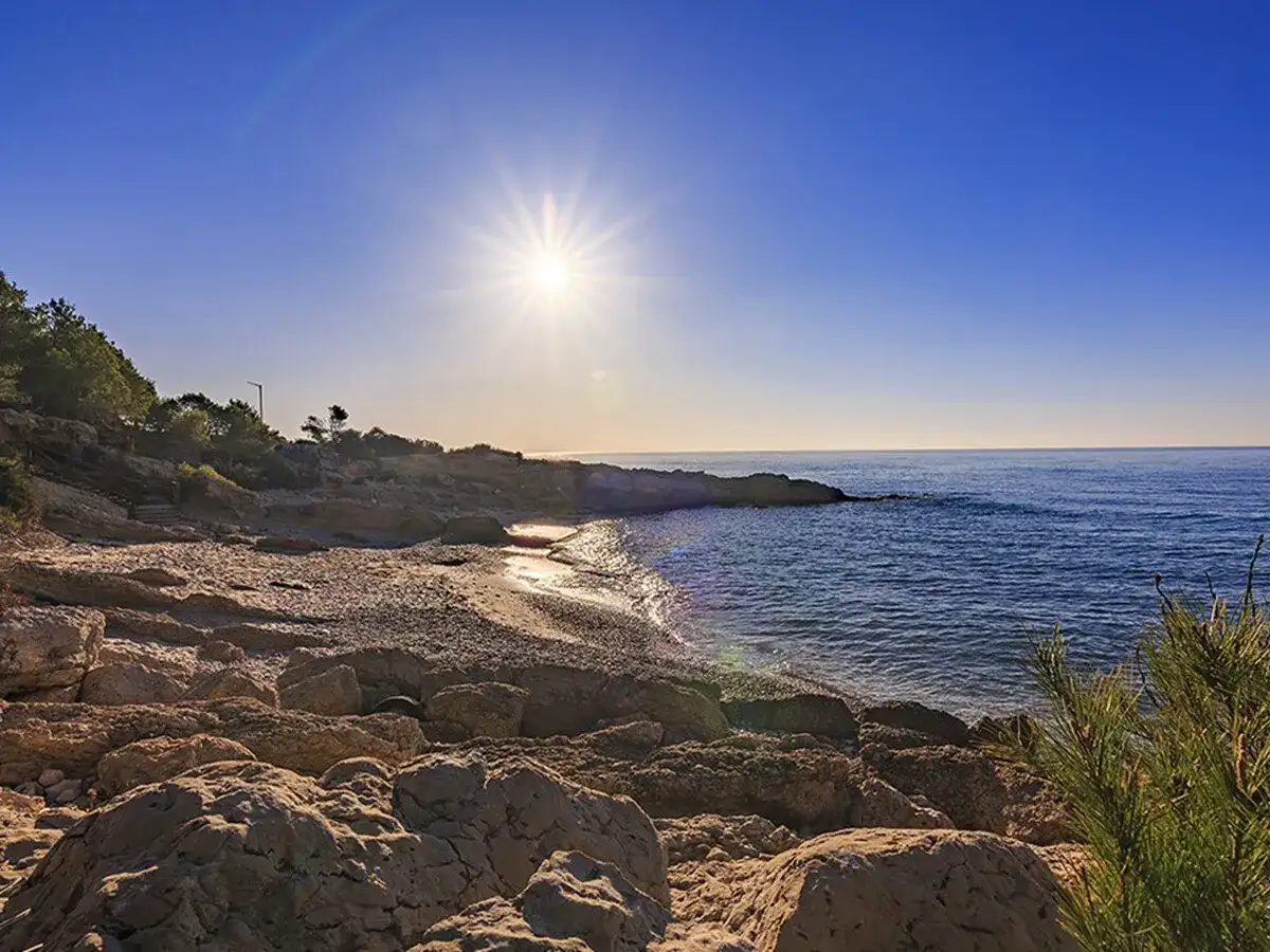 Sol radiante sobre una cala blanca de rocas y arena. El mar azul se extiende hasta el horizonte.
