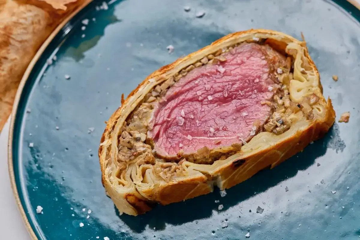 Un jugoso solomillo Wellington, envuelto en hojaldre dorado y champi&ntilde;ones, reposa en un plato azul.