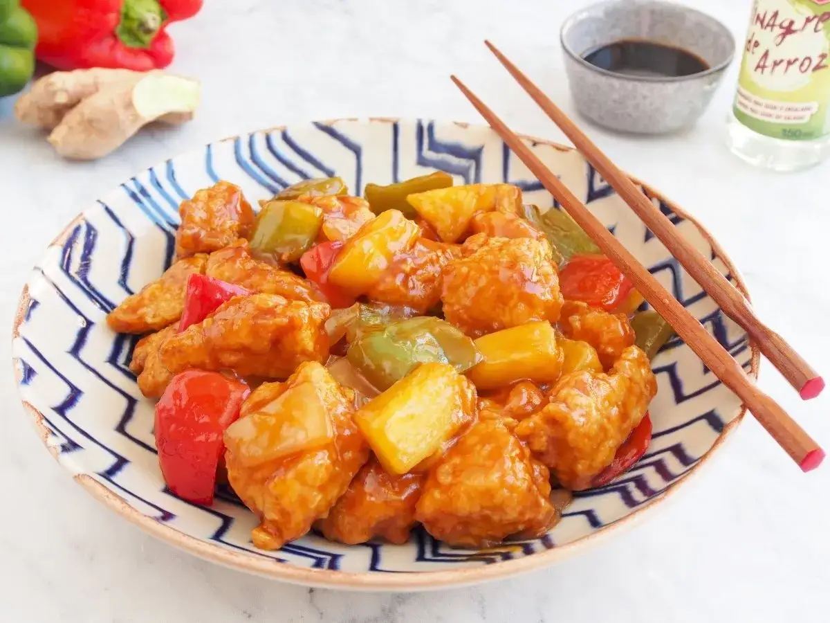 Delicioso cerdo agridulce con trozos de pi&ntilde;a y pimientos en una salsa brillante, servido en un plato decorado con palillos.