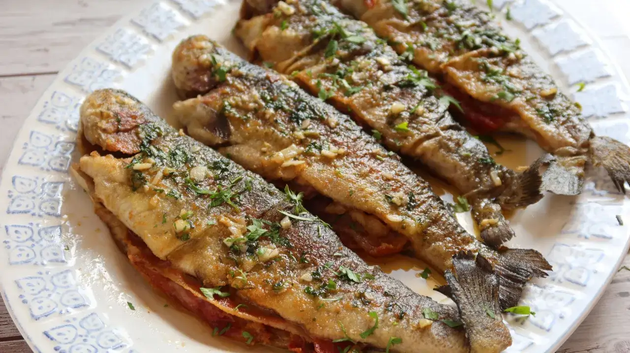 Cuatro truchas a la navarra doradas, rellenas y espolvoreadas con perejil fresco y ajo picado, servidas en un plato blanco con borde azul.