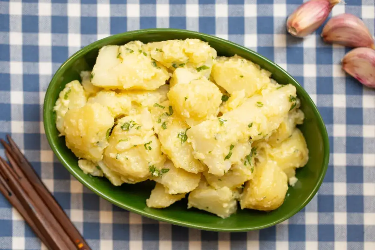 Un taz&oacute;n verde lleno de patatas alioli cremosas, espolvoreadas con perejil fresco. A su lado, unos dientes de ajo listos para m&aacute;s alioli.