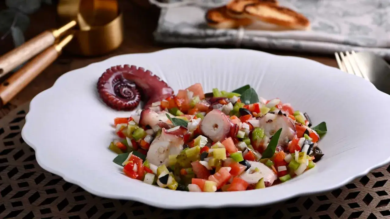 Delicioso salpic&oacute;n de pulpo con verduras frescas y aceitunas negras, servido en un plato blanco con un tent&aacute;culo de pulpo en espiral.