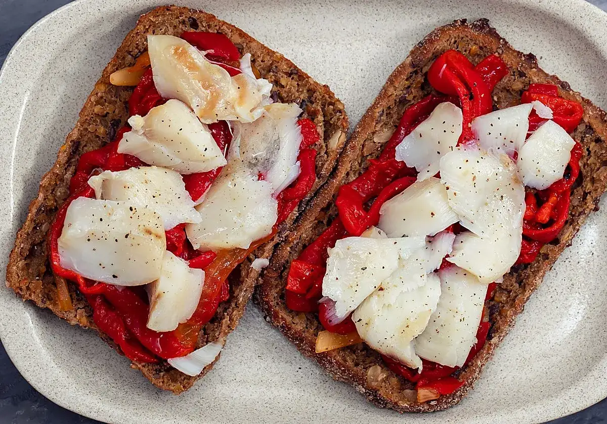 Dos tostadas de pan integral con pimientos rojos asados y trozos de brandada de bacalao, espolvoreadas con pimienta negra.