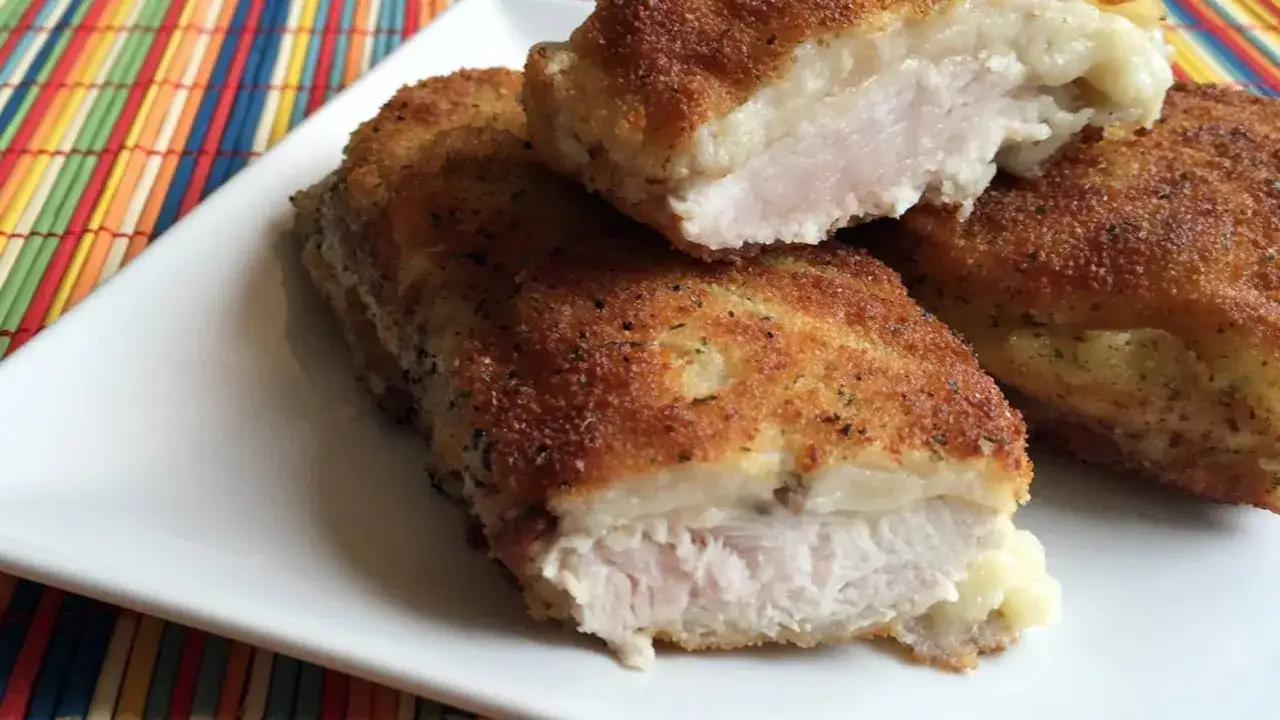 Pechuga villaroy dorada y crujiente, rellena de queso fundido, servida en un plato blanco sobre un mantel colorido.