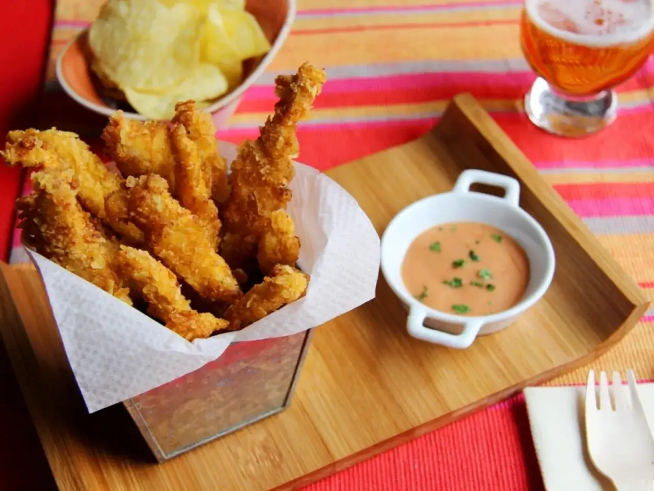 Crujientes tiras de pollo frito servidas en un cubo metálico, acompañadas de salsa rosa y patatas fritas.