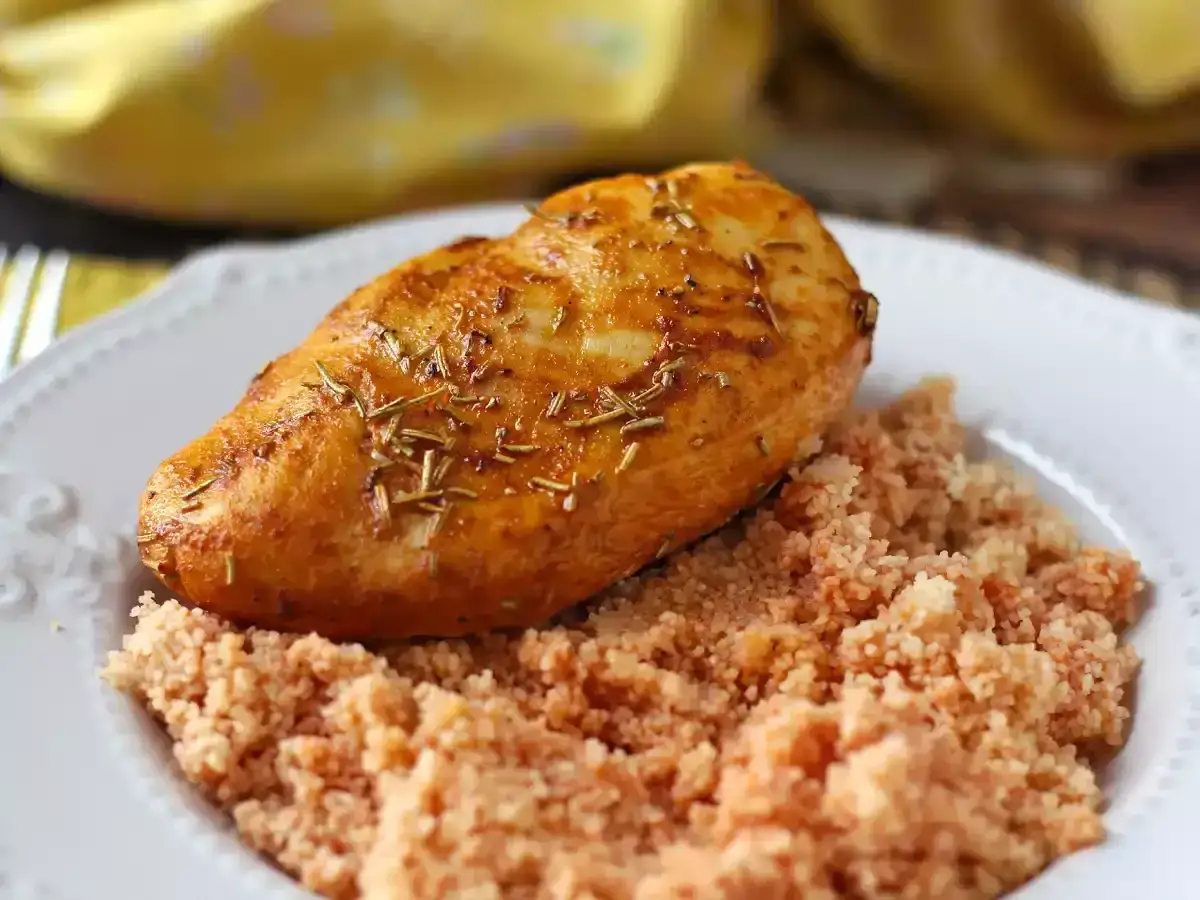 Pechuga de pollo dorada y jugosa, con hierbas arom&aacute;ticas, servida sobre una cama de cusc&uacute;s rosado en un plato blanco.
