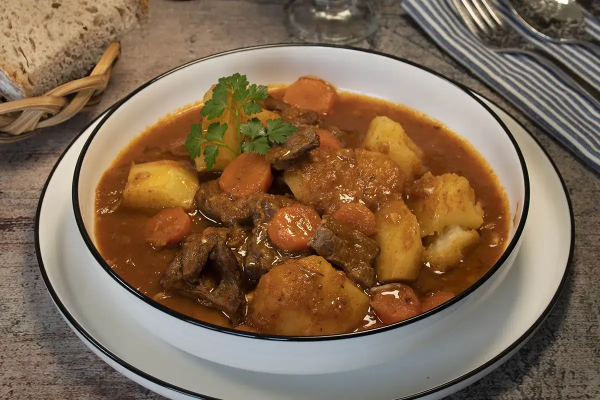 Un reconfortante ragout de ternera con patatas y zanahorias, servido en un cuenco blanco con pan r&uacute;stico al lado.