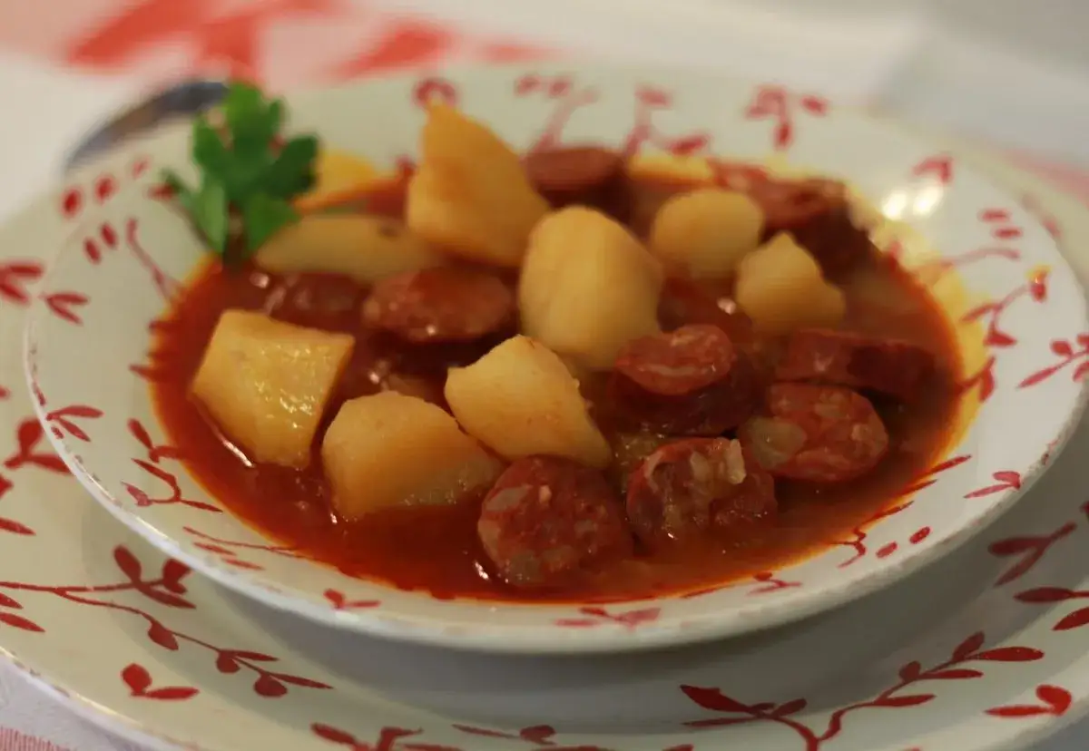 Un plato humeante de patatas a la riojana, con trozos de chorizo y patata en una salsa rojiza, adornado con una ramita de perejil.