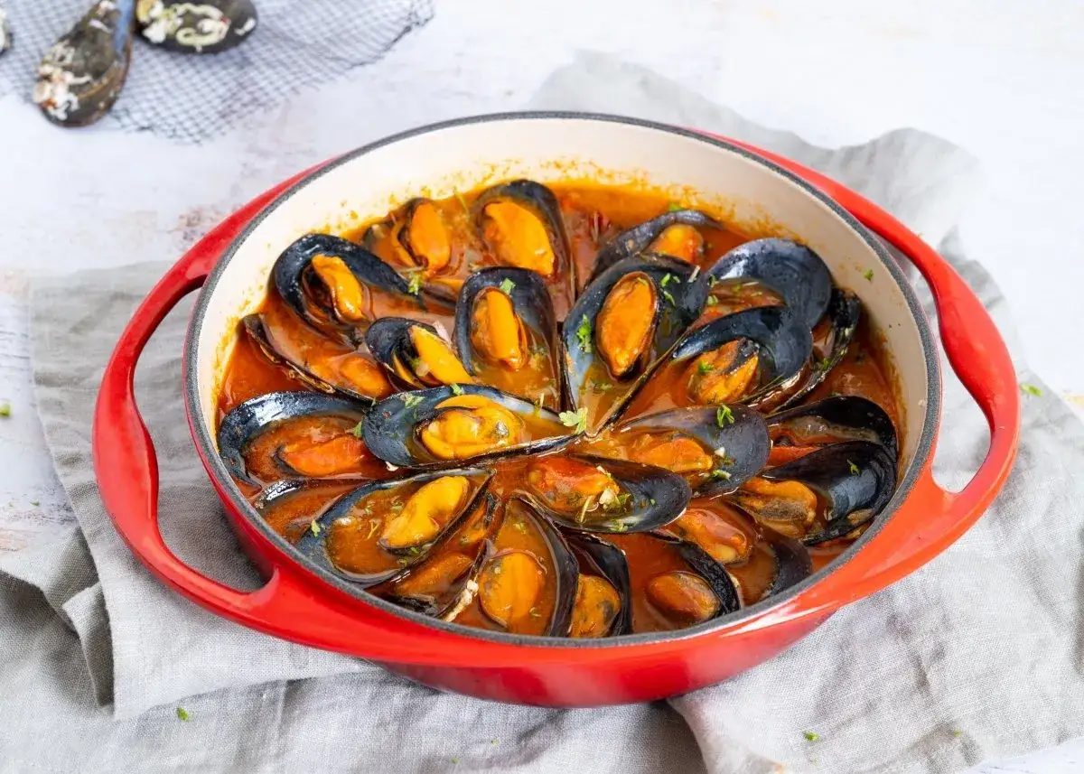 Mejillones a la marinera en una cazuela roja, con salsa sabrosa y perejil picado.