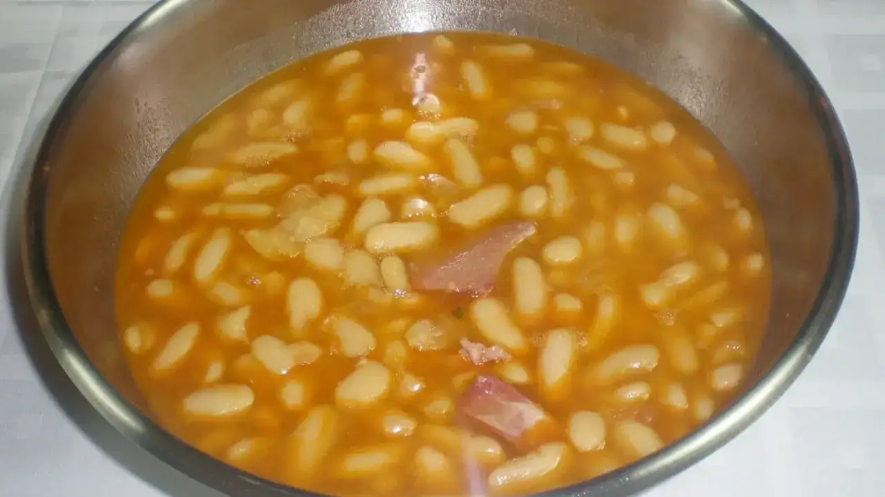 Un cuenco met&aacute;lico lleno de alubias blancas en un caldo anaranjado con trozos de jam&oacute;n.