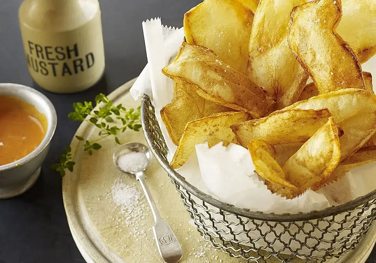 Patatas souffl&eacute; crujientes y doradas en una cesta de alambre, acompa&ntilde;adas de salsa y sal.