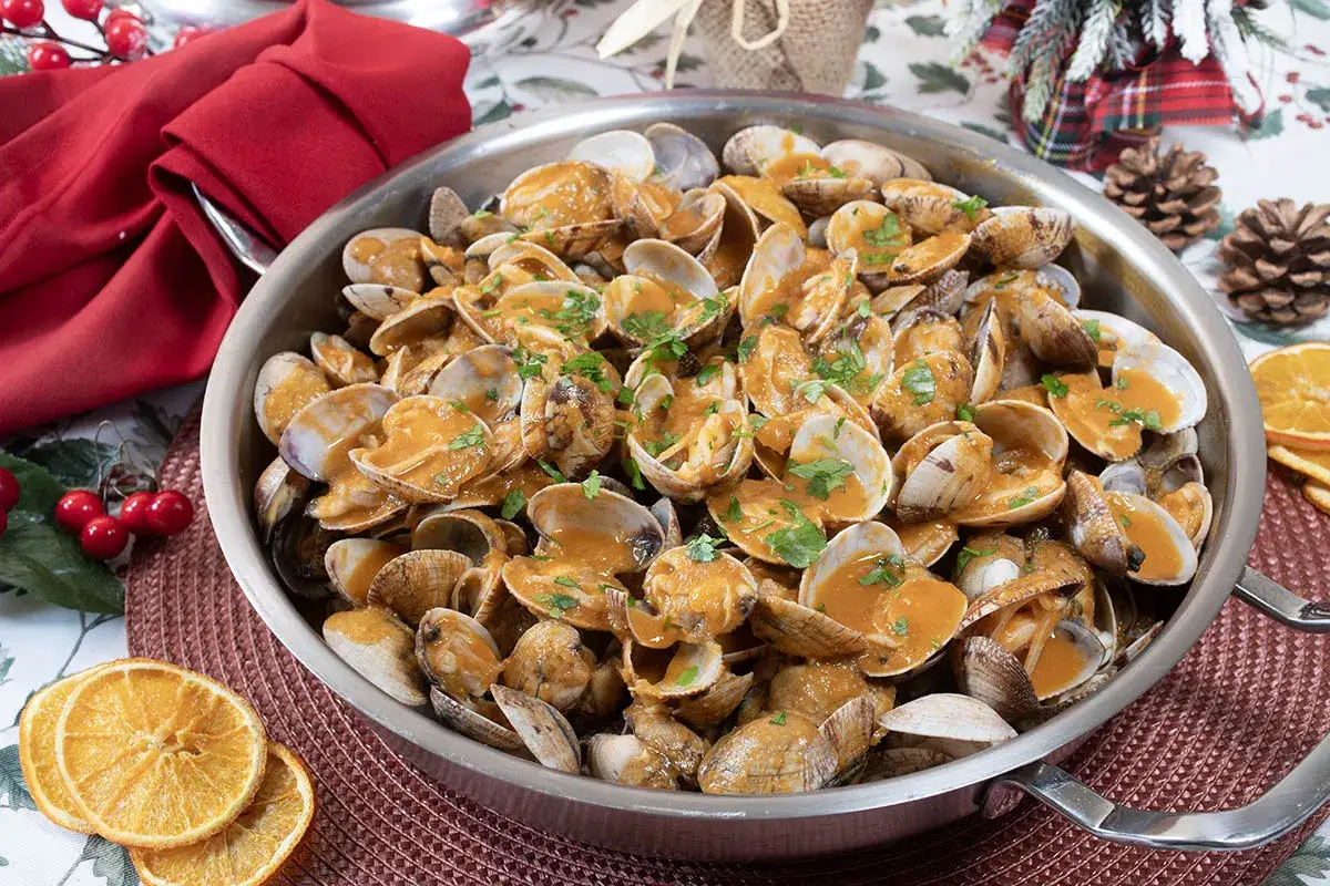 Un festín de almejas a la marinera, servidas en una sartén metálica, decoradas con perejil fresco y rodajas de naranja.