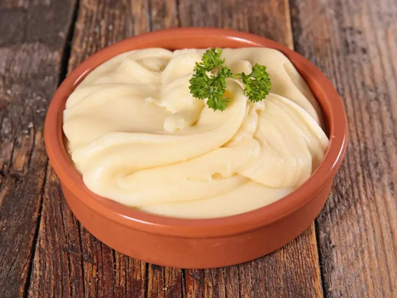 Un cuenco de barro con cremoso pur&eacute; de patatas, adornado con una ramita de perejil fresco, sobre una mesa de madera r&uacute;stica.