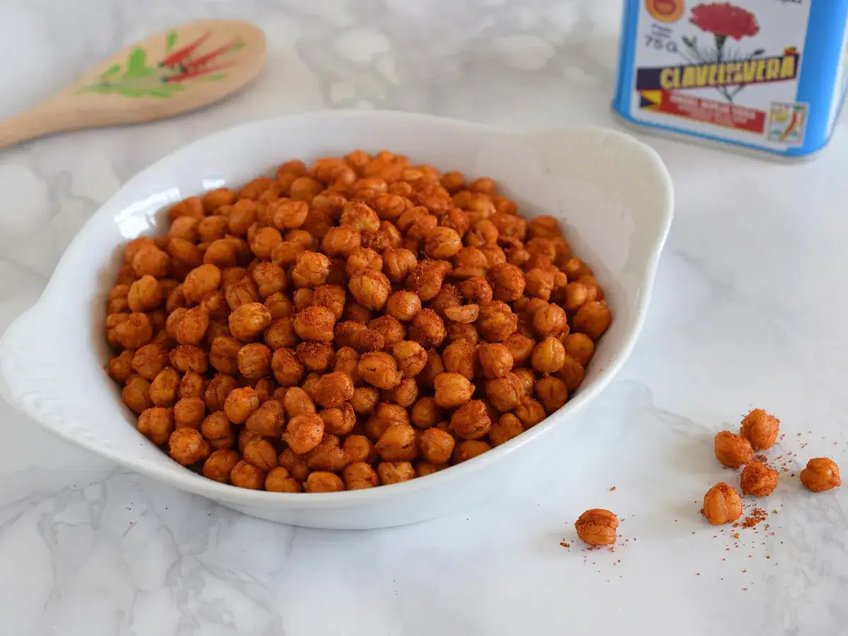 Un tazón blanco lleno de garbanzos fritos crujientes y especiados. Una lata de clavo y una cuchara de madera decorada están al fondo.