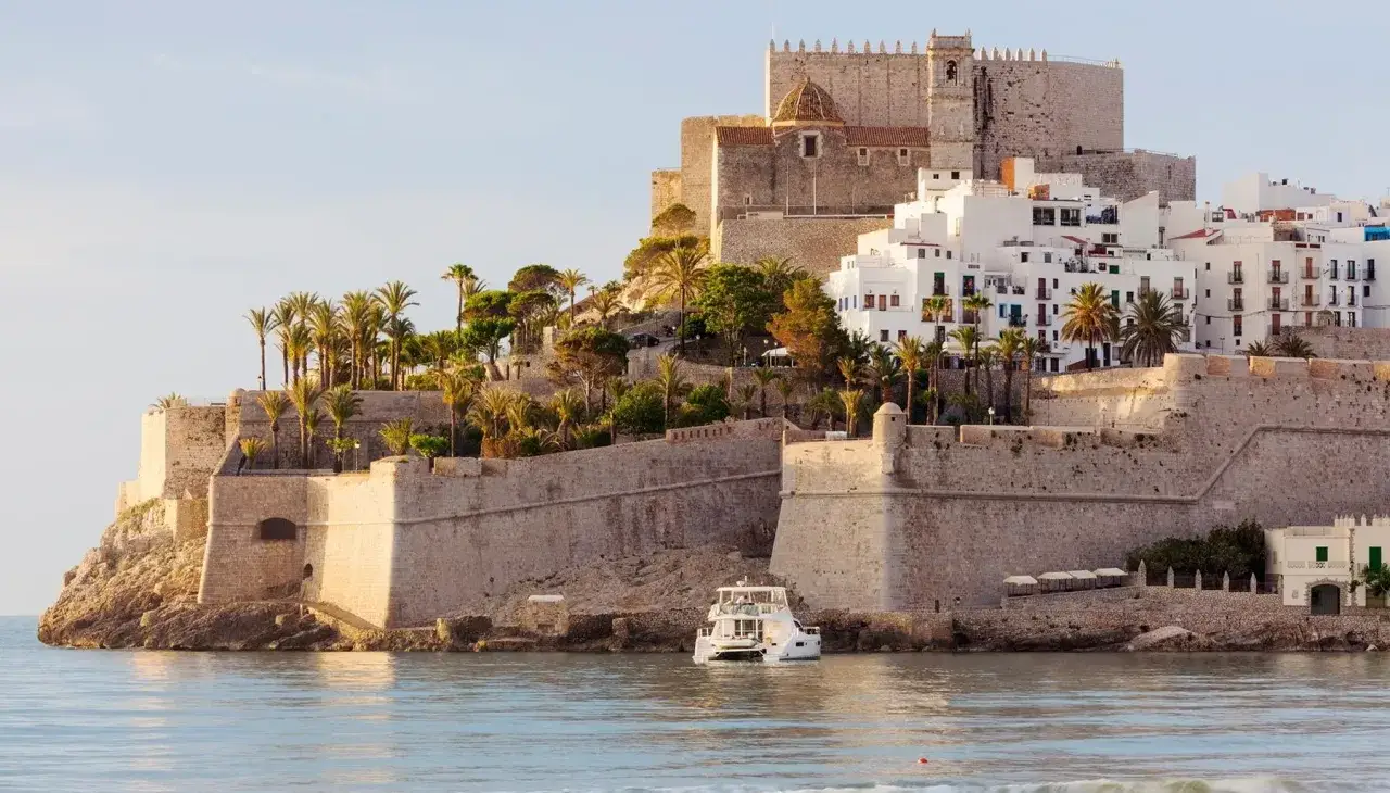 Peñíscola desde el mar, con su imponente castillo y casas blancas escalonadas en la colina. Un yate blanco navega tranquilamente.