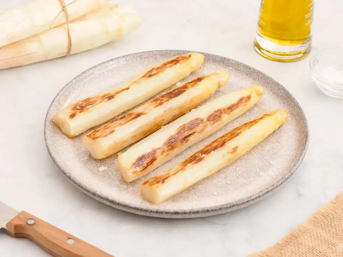 Cuatro espárragos blancos a la parrilla, con marcas doradas, reposan en un plato. Un manojo de espárragos blancos frescos yace al fondo.