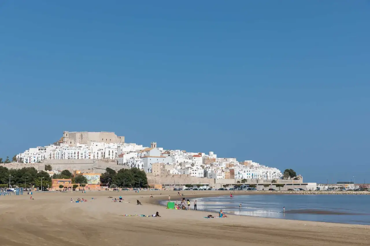 Gente disfruta del sol en la playa sur Pe&ntilde;&iacute;scola, con el castillo y el pueblo blanco al fondo.