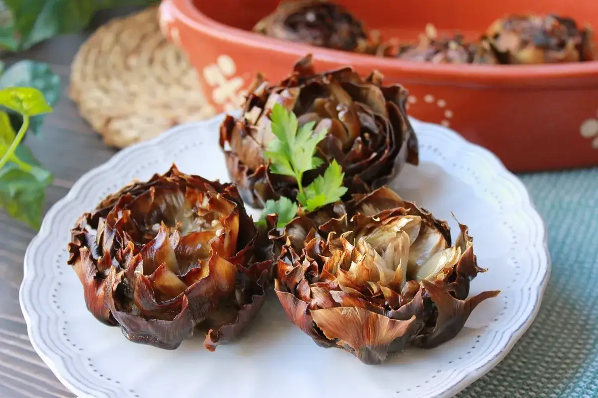 Tres alcachofas al horno, doradas y tiernas, servidas en un plato blanco con una ramita de perejil.