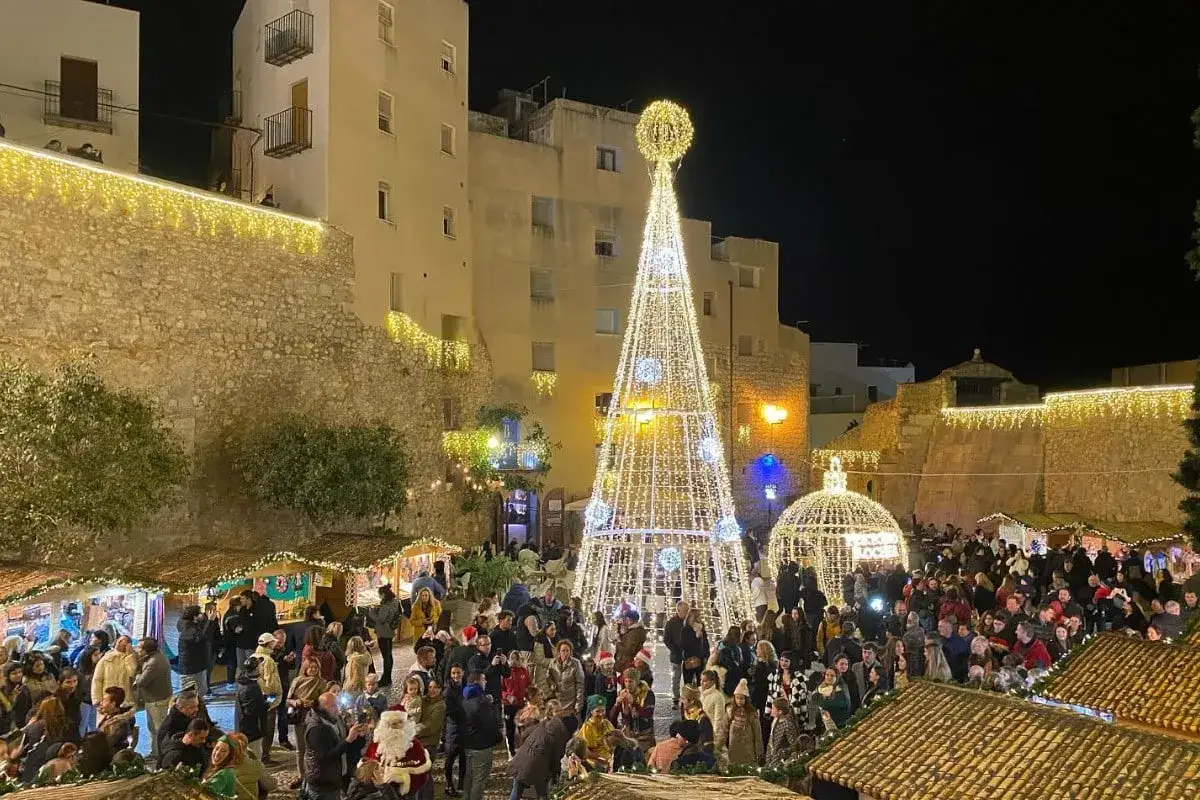Noche m&aacute;gica en el mercado navide&ntilde;o Pe&ntilde;&iacute;scola. Un &aacute;rbol gigante iluminado y puestos llenos de gente crean un ambiente festivo.