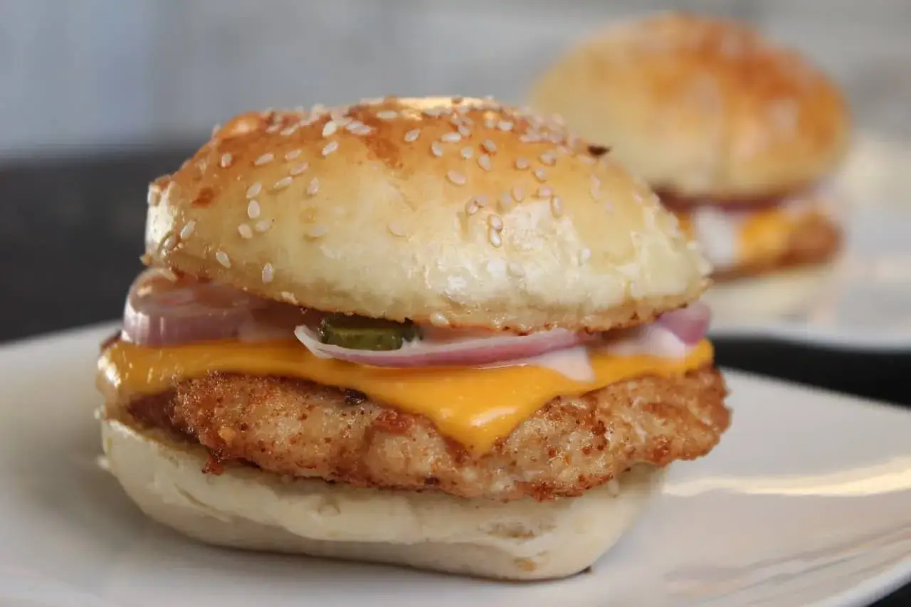 Deliciosa hamburguesa de pollo con queso derretido, cebolla morada y pepinillos, servida en un pan con semillas de sésamo.