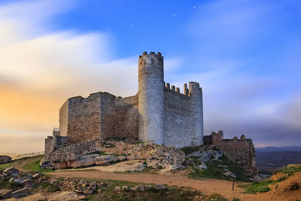 El imponente castillo de Xivert se alza sobre una colina bajo un cielo estrellado y nubes que se mueven, con tonos anaranjados al amanecer.