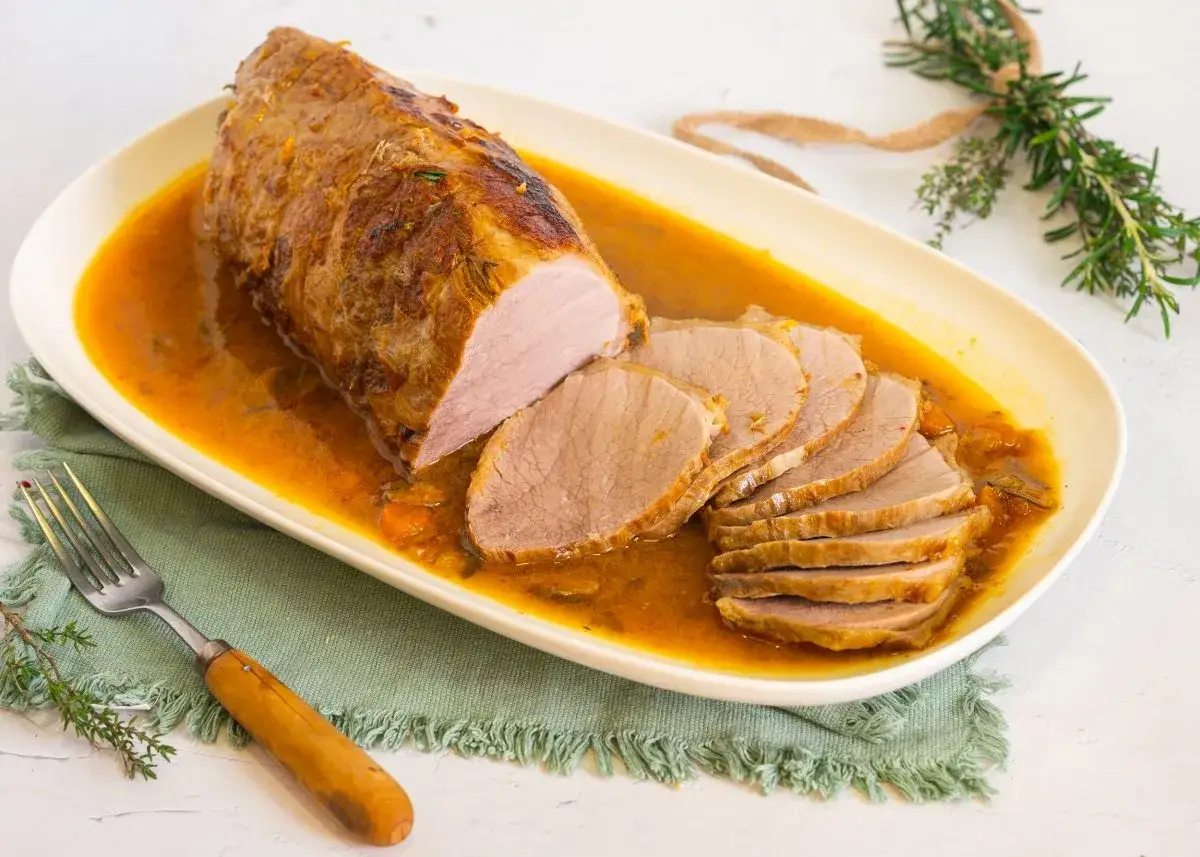Jugoso roast beef en salsa dorada, servido en una bandeja ovalada con rodajas tiernas y un tenedor r&uacute;stico.