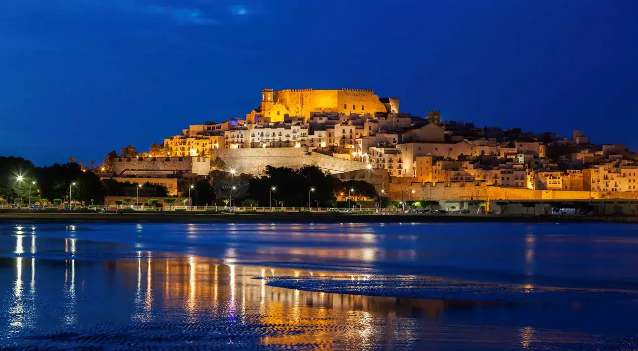 Noche estrellada sobre la playa sur Pe&ntilde;&iacute;scola, con el castillo iluminado y reflejos en el mar.