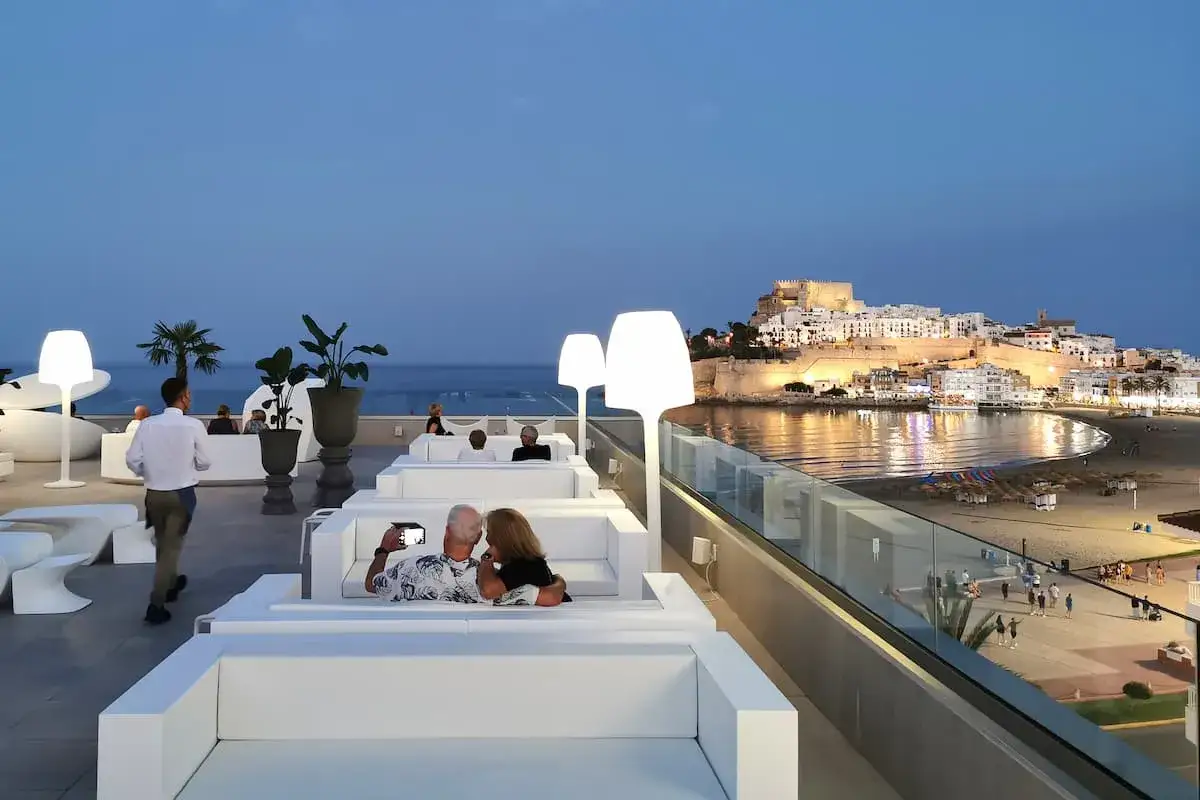 Pareja se toma selfie en el chiringuito de Pepe, con vistas a un pueblo blanco iluminado junto al mar.