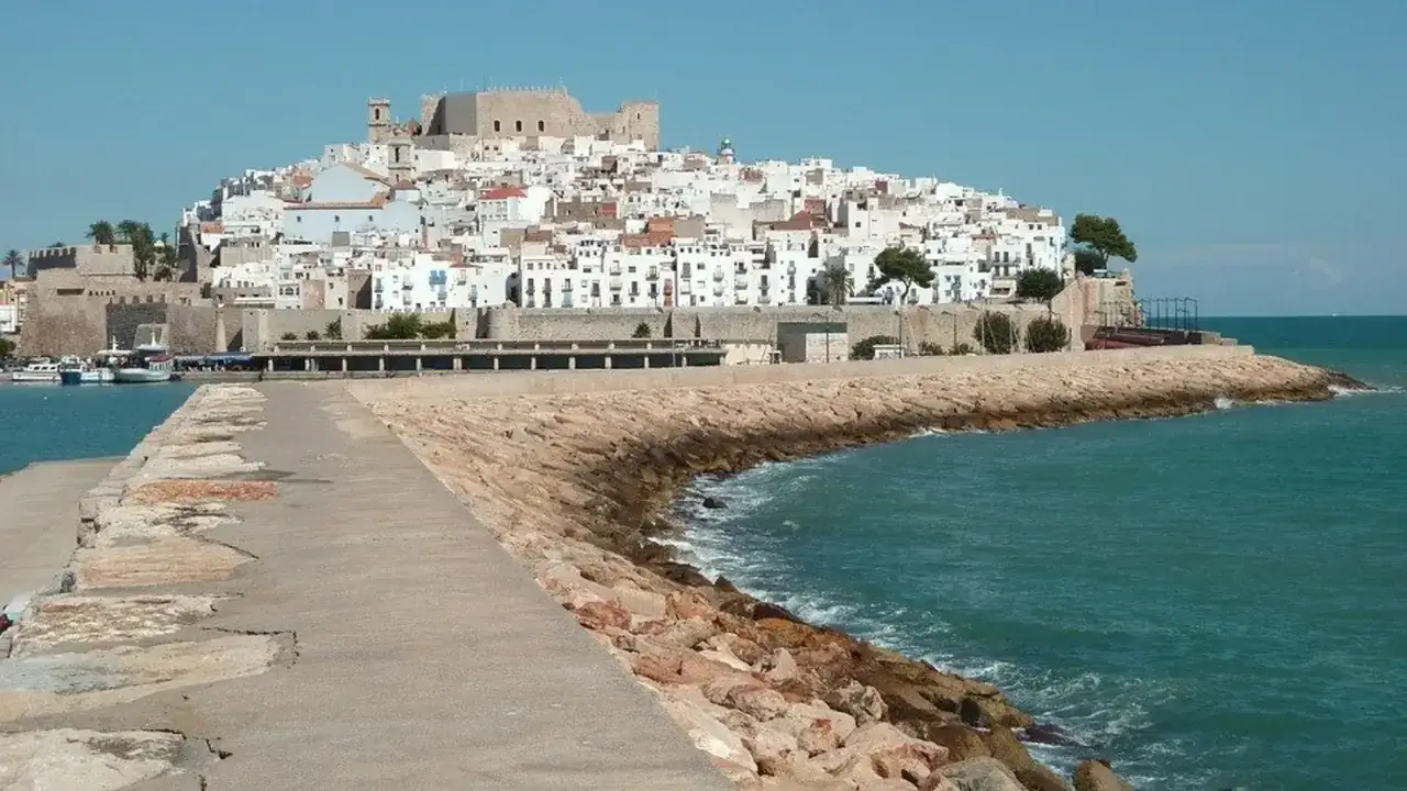 Pueblos costa Azahar: un castillo domina el caser&iacute;o blanco apilado en una colina, con el mar turquesa rompiendo contra un rompeolas de rocas.