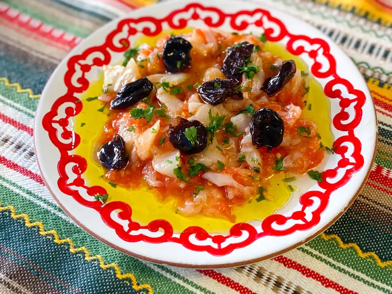 Deliciosa ensalada de bacalao con aceitunas negras y perejil, ba&ntilde;ada en aceite de oliva, servida en un plato blanco con borde rojo.