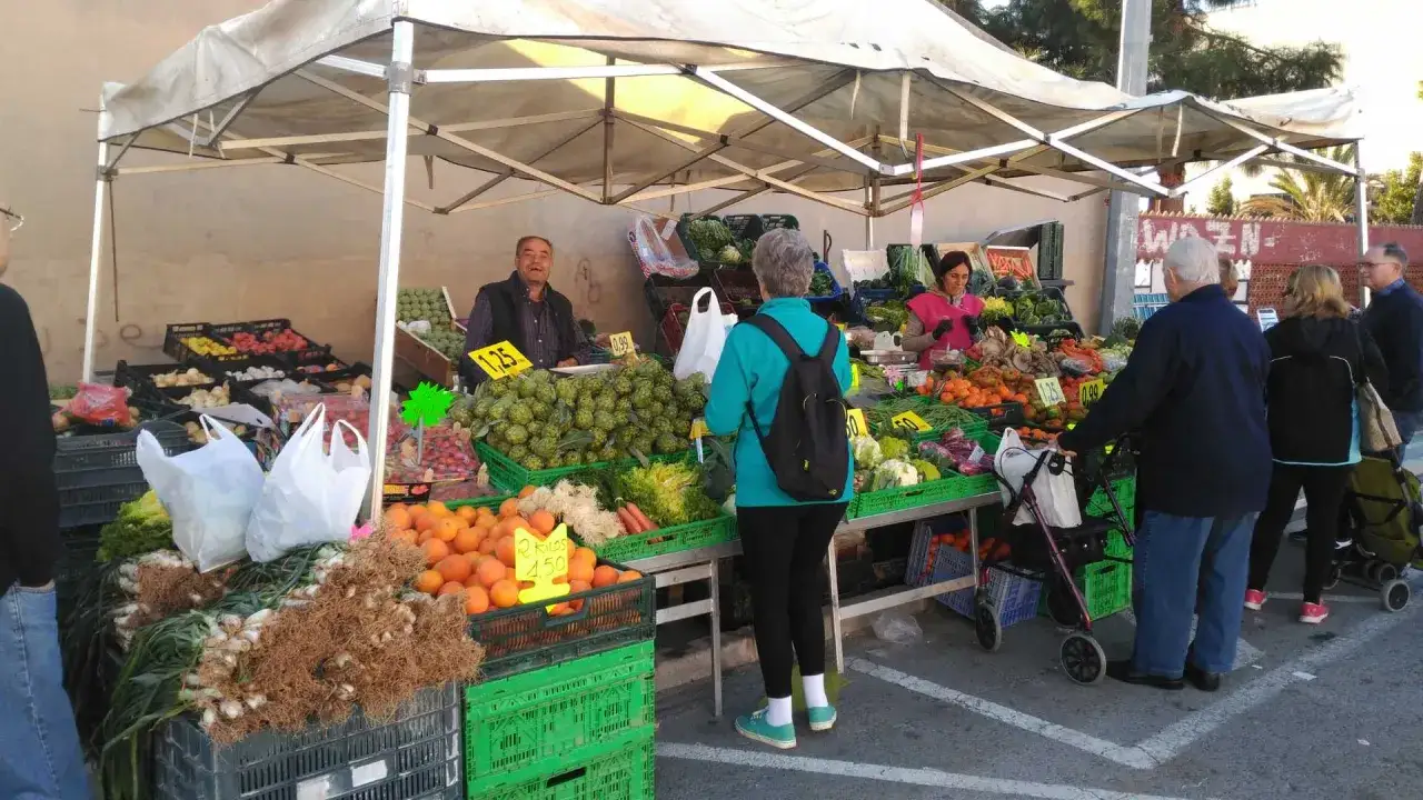 Mercadillo en Benic&agrave;ssim con puestos de frutas y verduras frescas. Un hombre sonriente atiende a los clientes.