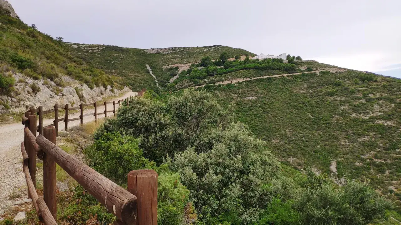 Sendero de montaña con vallas de madera, vegetación exuberante y edificios blancos en la cima. Un tramo del camino de Santiago Peñíscola.