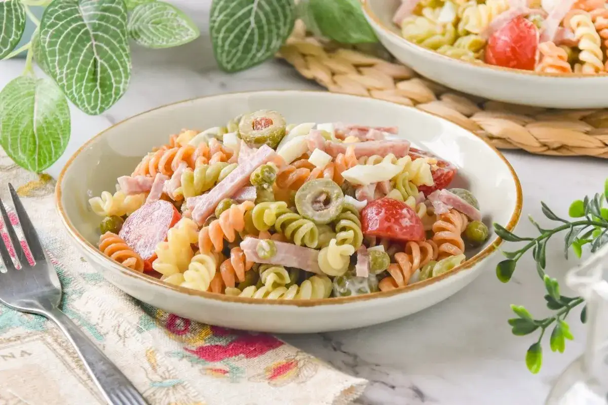 Un plato de ensalada de pasta con trozos de jamón, huevo duro, aceitunas y tomates cherry.
